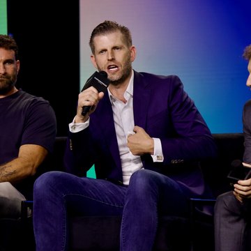 Eric Trump, wearing a purple blazer and white shirt, speaking into a microphone while seated on a stage panel.