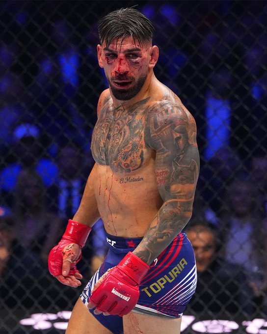 The image features a man with tattoos, wearing boxing gloves and standing in front of a crowd at an event. The man appears to be bruised or injured, possibly from a fight. He is holding a pair of boxing gloves, which suggests that he might have been participating in a match or training session. The post about the fighter's return after being out sick by UFC has a connection to this image as it highlights the fighter's determination and resilience despite his injuries.