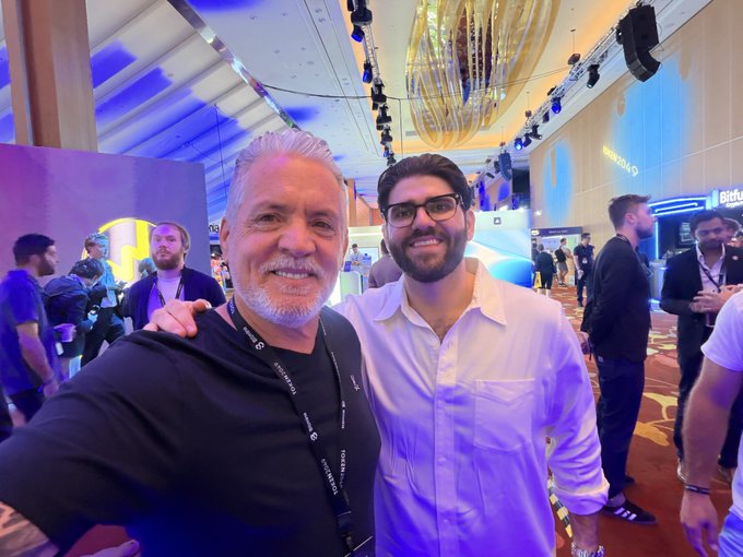 Two men posing for a photo at a conference or event venue with blue lighting and a Bitcoin logo visible in the background.