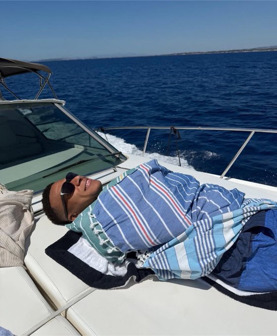 A man wearing sunglasses relaxes on the deck of a moving boat, wrapped in a blue striped towel, with the open blue sea stretching to the horizon.