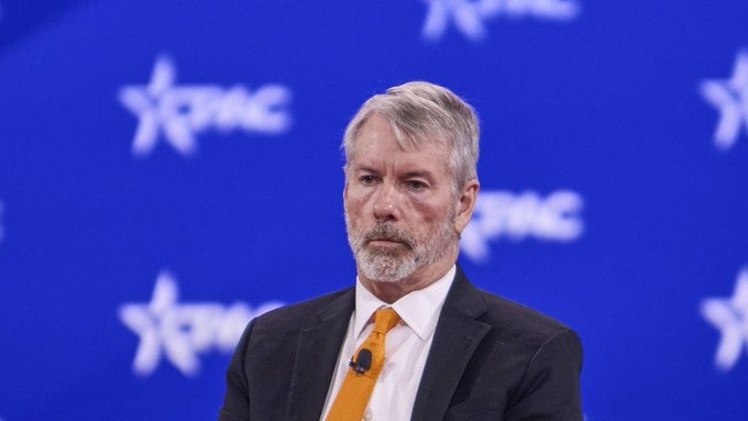A man with gray hair and a beard, wearing a dark suit with a white shirt and orange tie, speaks at a CPAC event with the blue branded backdrop behind him.