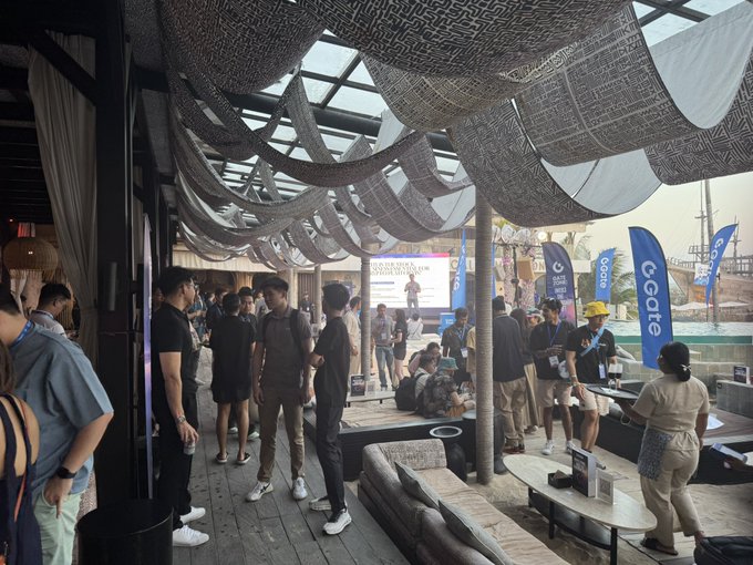 Photograph of a crowded outdoor lounge area at what appears to be a Gate.io-sponsored event near a beach, with attendees mingling under fabric canopy drapes and blue Gate promotional flags.