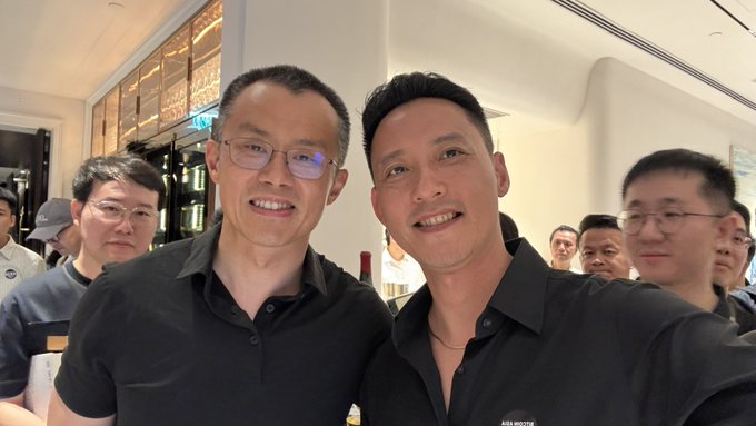 Two men pose for a photo at what appears to be a social gathering, with one wearing a black polo shirt and glasses, and the other in a dark shirt. The context suggests this is a meeting between Zhao Changpeng (CZ), founder of Binance, and his wife He Yi (何一), who is also a co-founder of Binance, capturing a personal moment between the prominent crypto entrepreneurs.