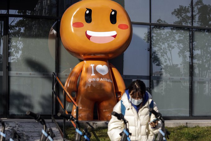 A large orange inflatable mascot wearing an 'I ❤️ QIN@Alibaba' shirt stands outside a glass building while a masked person in a white jacket walks by with bicycles in the foreground.
