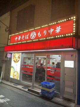 The image shows a restaurant with an Asian-style sign, indicating that it serves Asian cuisine. The exterior of the building features a red awning over the entrance, which adds color and character to the establishment. A blue trash can is located near the entrance, likely for waste disposal purposes. There are several people inside the restaurant, suggesting that it is currently in use or about to open for business.