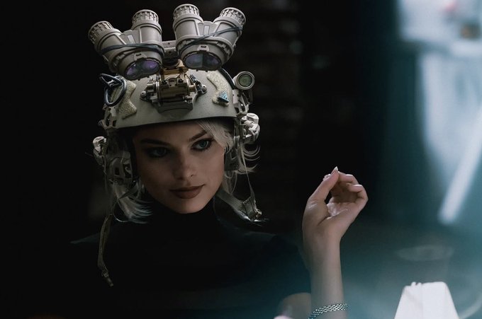A cinematic portrait of a woman with blonde hair wearing an elaborate, futuristic helmet adorned with mechanical eyes and lenses, set against a dark background.