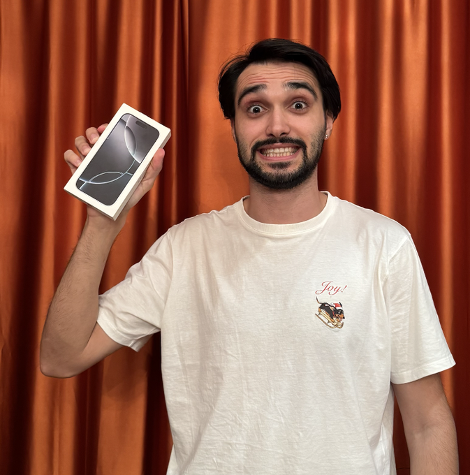 A bearded man wearing a white t-shirt with a small graphic stands in front of an orange curtain, holding up a white box containing a dark smartphone with an excited expression.