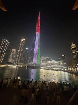 A nighttime view of the Burj Khalifa illuminated in red, green, and purple colors, seen from across a plaza where people are gathered.