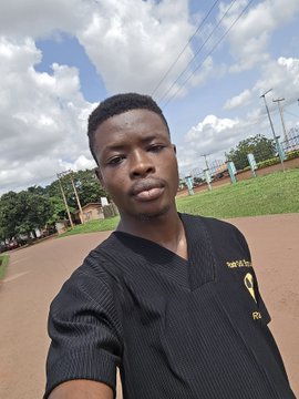 A young man wearing a black shirt with yellow text takes a selfie outdoors on a paved path with a grassy field, trees, and cloudy sky in the background.