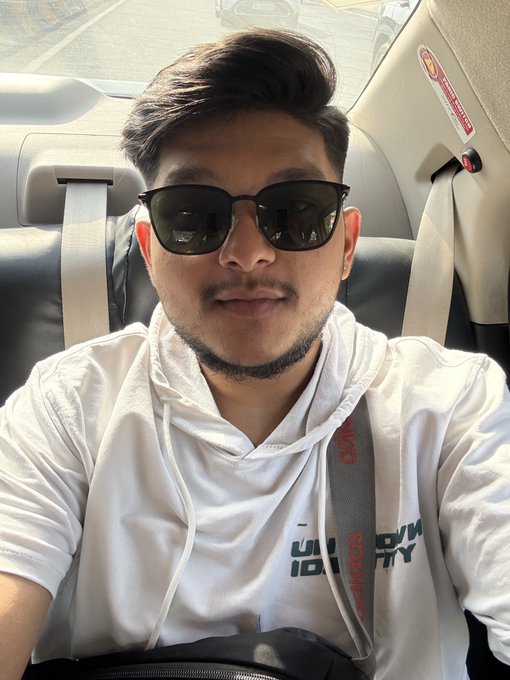 A selfie of a young man with dark hair and a beard wearing black sunglasses and a white hoodie, sitting in the backseat of a car.