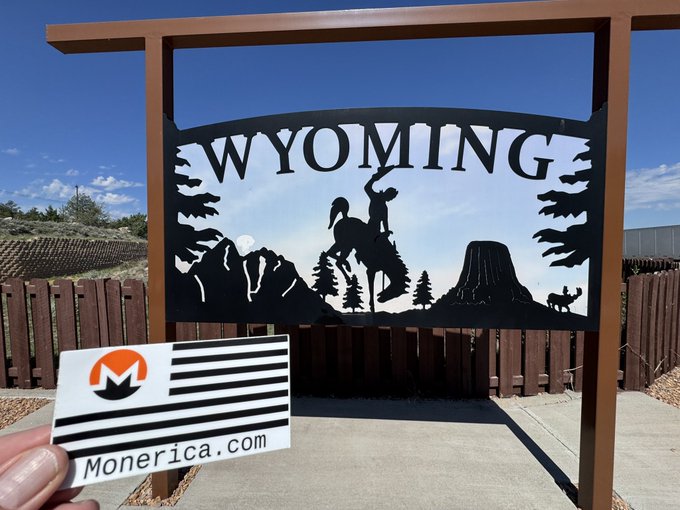 A metal cutout sign reading 'WYOMING' with a cowboy and wildlife silhouette, held in front of a fenced area with a Monerica.com business card.