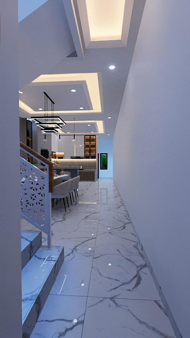 A modern interior hallway features white marble flooring with grey veining, a recessed ceiling with warm lighting, a staircase with a decorative white railing on the left, and a kitchen area with a dining table in the background.