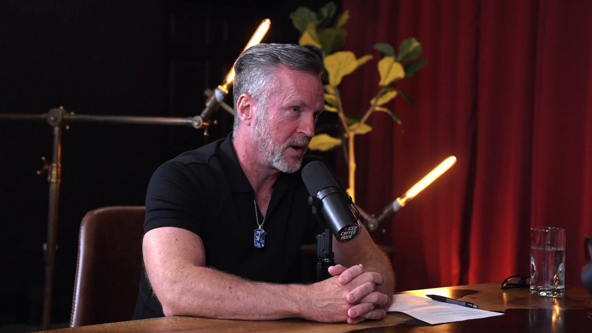 A man sits at a desk with his hands clasped together while holding a microphone. The background features a red curtain, indicating that he may be in an indoor setting or at a conference. He appears to be wearing a black t-shirt as well. In front of him on the desk is a cup filled with water. The man's words are related to his personal finances, specifically focusing on the value of his properties and expenses like rent or car payment. The image also contains some text that reads "Michael Zuber reveals he owns 167 units and about 25 million in total value" which might be providing further context to his financial situation.