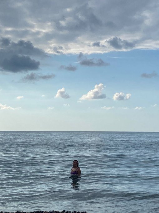 A person stands chest-deep in calm ocean water under a bright blue sky scattered with clouds.
