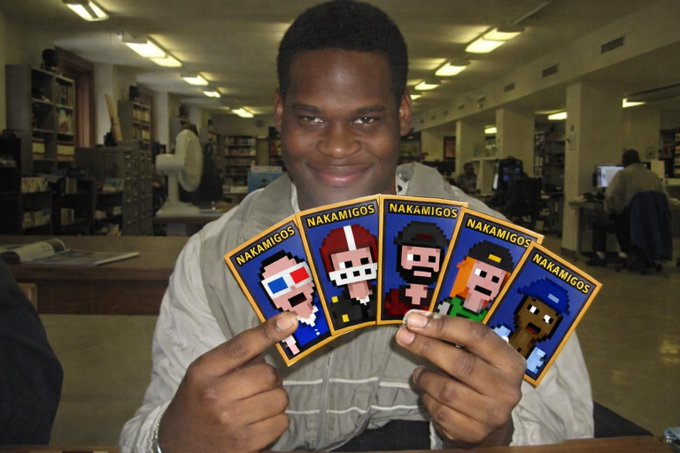 A smiling young man in a library setting holds up five pixel-art character cards labeled 'NAKAMIGOS' featuring various colorful figures.