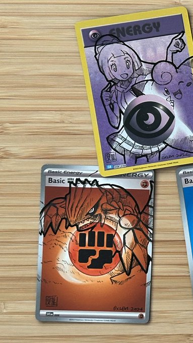 A photograph of custom Pokémon trading cards on a wooden surface, featuring hand-drawn Psychic Energy and Basic Fire Energy cards with original artwork signed 'Julem 2025'.