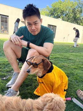 The image features a man sitting on grass with his dog wearing an orange raincoat, both of them smiling for the camera. The background shows other people walking around outside, indicating that they are likely at a park or similar outdoor location. There is also a handbag visible in the scene, suggesting that someone might have brought it along to enjoy some time outdoors with their pet and friends.