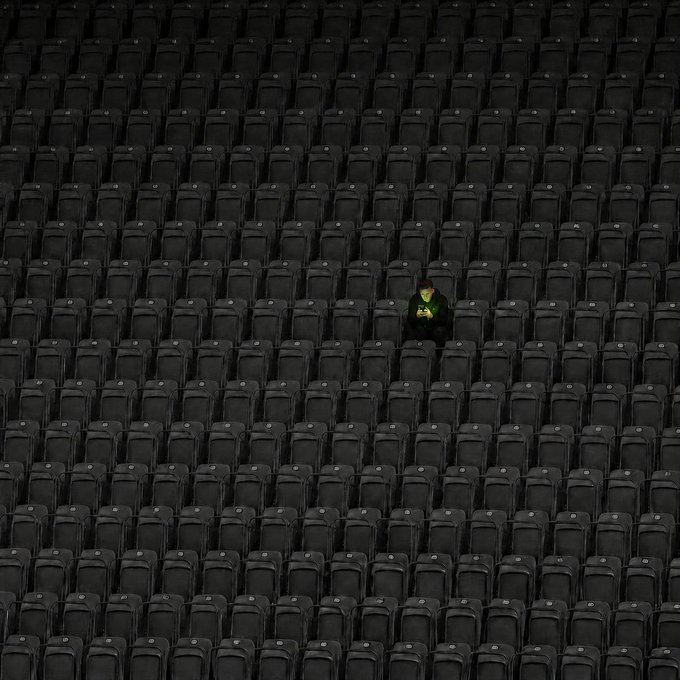 A lone person sits amid rows of empty stadium seats, their yellow jacket standing out against the sea of black chairs. The image visually echoes the message of solitary early arrival—being alone not because of exclusion, but because they beat everyone else there.