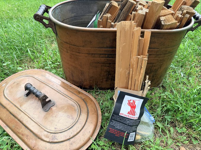 An old metal pot filled with chopped firewood sits on grass, with its lid beside it and a 'FreeSampah' flyer attached to the wood.