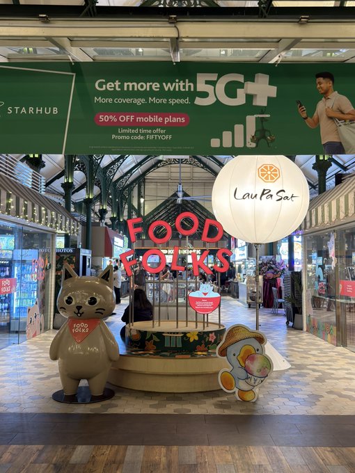 A promotional display at Lau Pa Sat food market in Singapore features a large "FOOD FOLKS" sign with cartoon mascots, while a StarHub banner above advertises "Get more with 5G+" and "50% OFF mobile plans" with promo code FIFTYOFF. The "Food Folks" branding ties into the caption's discovery of local cuisine, complementing the visit to this iconic Singapore food destination known for its Malay satay.