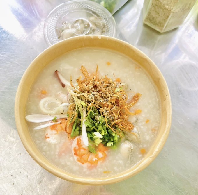 A bowl of congee or rice porridge is topped with crispy fried shallots, green onions, and a cooked shrimp, sitting on a metallic surface next to a small dish of pickled vegetables.