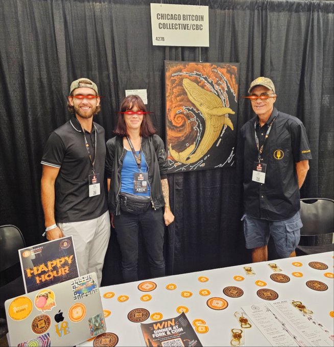 Three people wearing red-tinted glasses stand behind a booth for 'Chicago Bitcoin Collective/CBC' at a convention, with a whale artwork displayed behind them and Bitcoin memorabilia on the table.