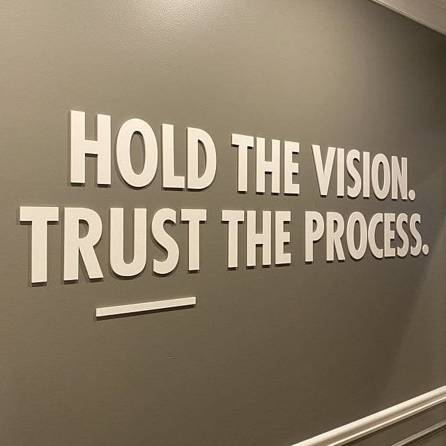 White 3D lettering on a gray wall displays the motivational phrase 'HOLD THE VISION. TRUST THE PROCESS.'
