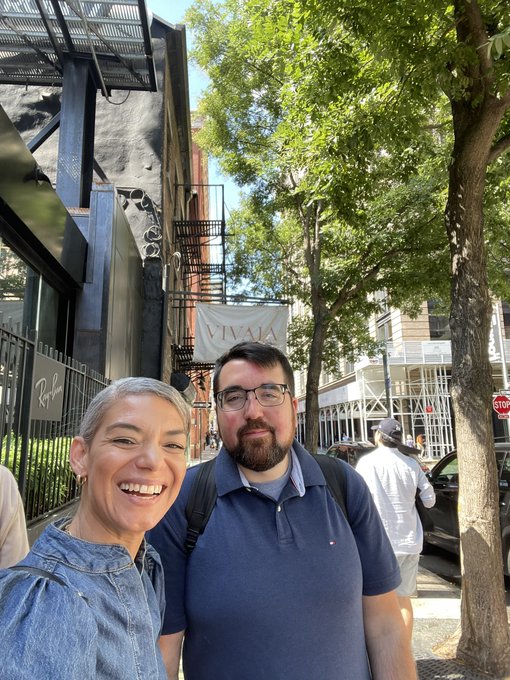 A smiling couple takes a selfie on a city street with a 'VIVALA' sign visible on a building behind them.