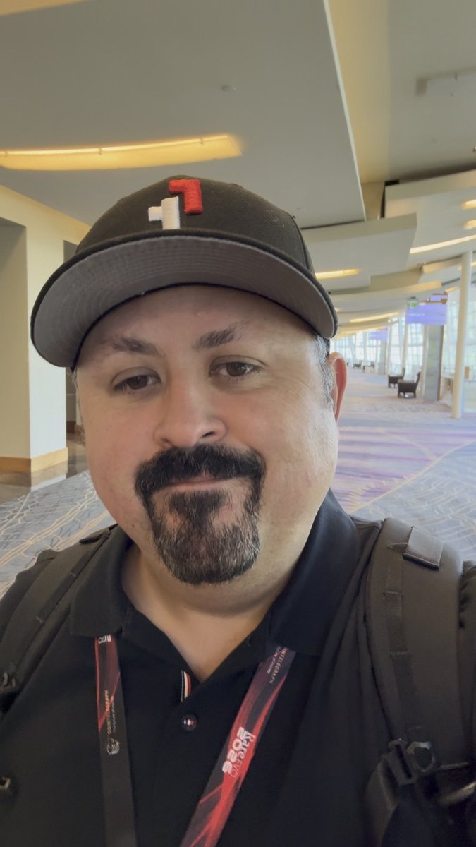 A selfie of a man with a goatee wearing a black cap with a red and white logo and a black shirt with a lanyard, taken inside what appears to be a convention center.