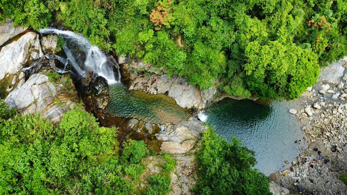 An aerial drone shot captures a waterfall cascading into a deep blue natural pool, surrounded by lush green forest and grey rocks.