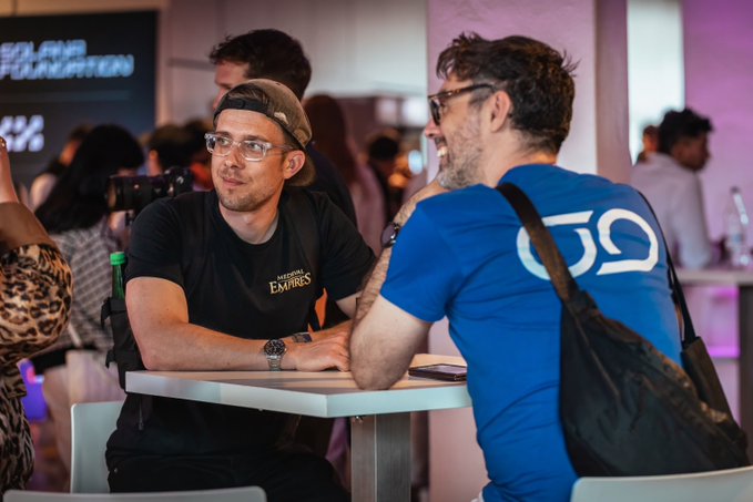 The image shows a group of people sitting around a table, engaged in conversation. There are two men prominently featured in the scene - one wearing a black shirt with "Emirates" written on it, and another man wearing a blue shirt. The other individuals present include a person holding a camera and a handbag.

The background reveals that this gathering is taking place at an event or conference, as indicated by the text "Conference maxxxing this week. Next stop: @Ronin_Network meet up on Thursday." This suggests that the group of people in the image might be attending a conference related to travel or transportation, possibly discussing topics such as airlines, travel experiences, or industry trends.