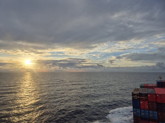 A photograph taken from a cruise ship or large vessel at sea during sunset, showing a colorful cloudy sky, sun reflecting on the ocean water, and stacked shipping containers on the right side.