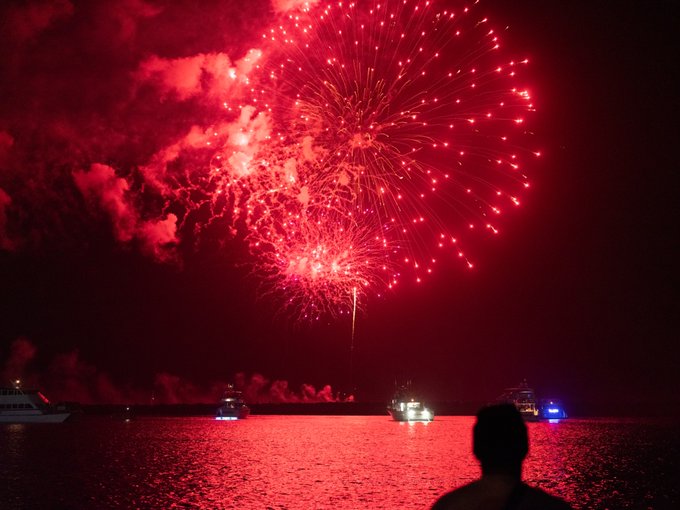 A vibrant red fireworks display over the ocean at night, with boats silhouetted on the water and a person watching in the foreground.