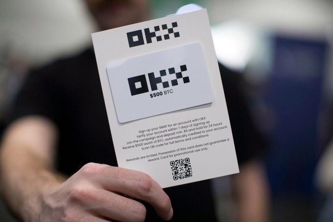 A person holds a promotional card for 'OKX' offering a '$500 BTC' reward for signing up and signing in, complete with a QR code.