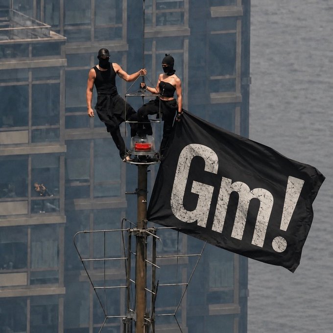 Two people wearing black masks and black clothing standing on a tall structure, holding up a large black flag with 'Gm!' written in white text.