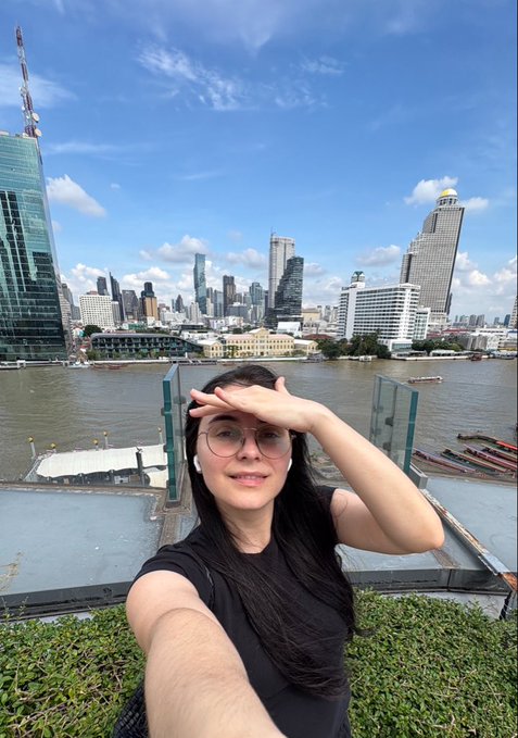 A woman in a black shirt and glasses takes a selfie in front of a city skyline with a river and boats visible in the background.