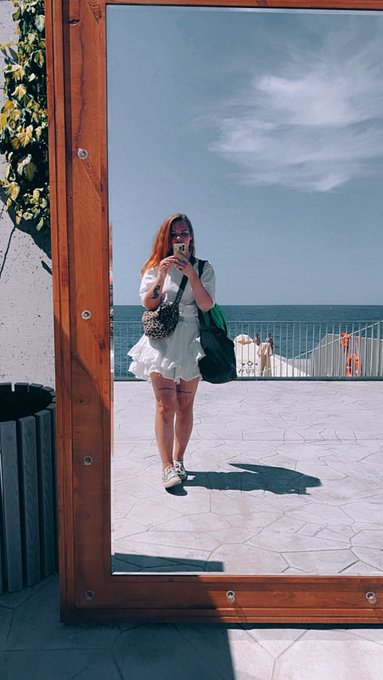 A woman in a white dress takes a mirror selfie on a seaside terrace, capturing herself against a bright blue sky and ocean horizon. The polished wooden-framed mirror serves as the "artwork" referenced in the post, titled "Sweet danger," offered as the final piece (3/3) of a collection for 40 XTZ on objkt.com.