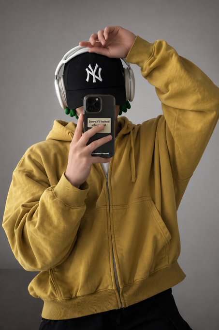 A person taking a mirror selfie wearing a yellow zip-up hoodie, a black NY Yankees cap, and silver headphones, holding a phone with a phone grip that says 'Sorry if I looked...'