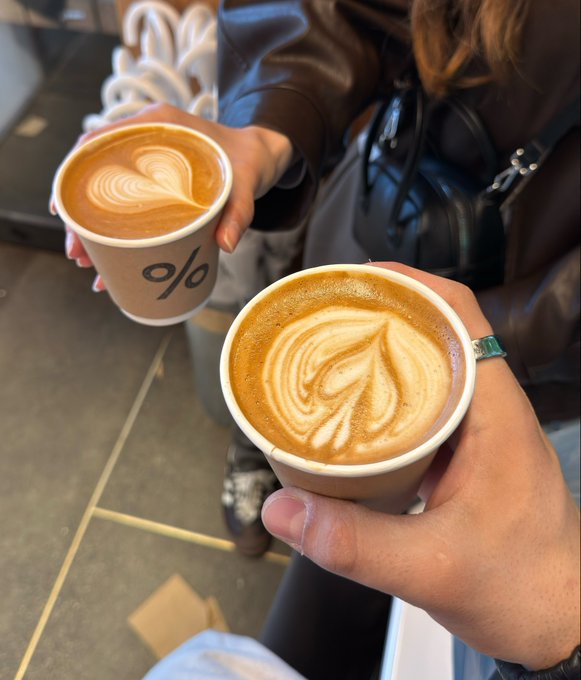 Two people holding coffee cups from '% Arabica' with latte designs on top, photographed from above on a tiled floor.