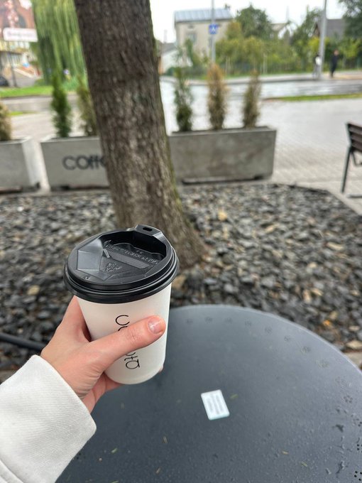 A hand holding a coffee cup with a black lid at an outdoor cafe, with a tree and planters visible in the background.