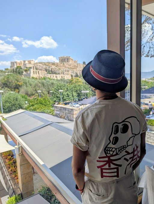 A person wearing a navy hat and a beige t-shirt with a skull design and Japanese characters, standing on a balcony overlooking the Acropolis in Athens, Greece.