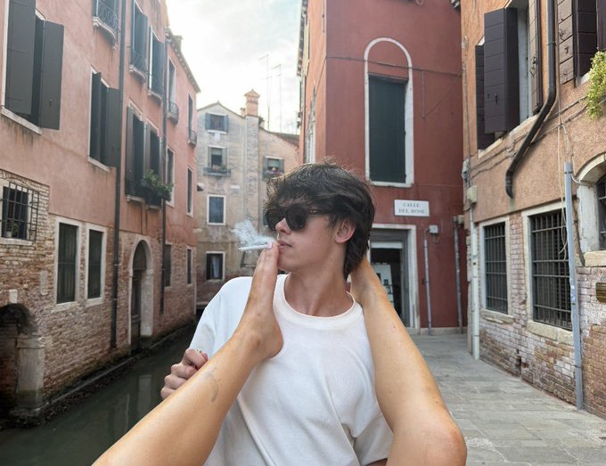 A young man in sunglasses smokes a cigarette while posing in a narrow Venice alleyway, with another person's hands playfully framing his face. The relaxed, tourist-like setting contrasts with the post's bold prediction about the subject's future fame surpassing SteveWillDoit's within two years, as well as their loyalty to Toshi.