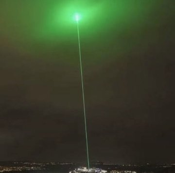 A green laser beam shoots vertically into a dark night sky from the distant horizon, where city lights are visible at the bottom of the frame.