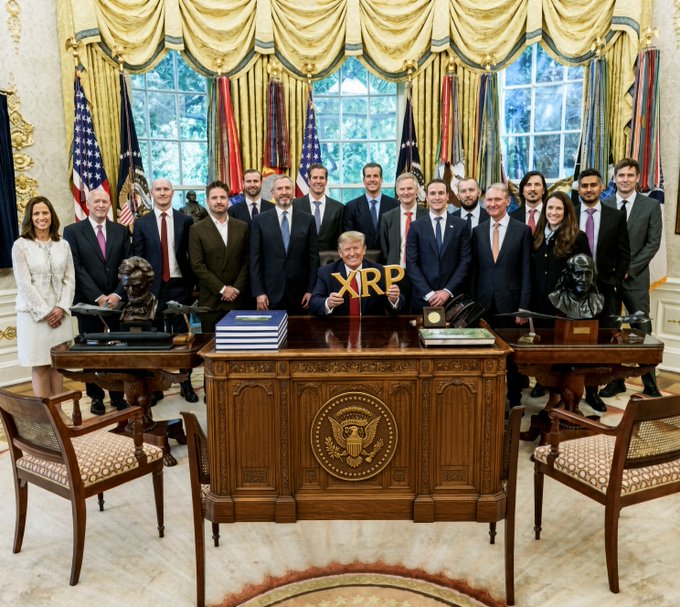 A group photo taken inside the Oval Office shows the President seated at the Resolute Desk surrounded by officials and tech executives, with an "XRP" sign prominently displayed on the desk in front of him—likely a playful reference tying the meeting to the XRP community's "XRPTrump" account mentioned in the post.