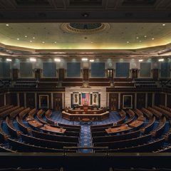 An empty congressional chamber or legislative hall with curved rows of blue seating facing a central podium, with a Brazilian flag visible on the right.