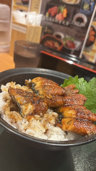 A close-up photo of a bowl of rice topped with grilled eel (unagi) and garnished with a shiso leaf, set in a restaurant with a menu visible in the background.