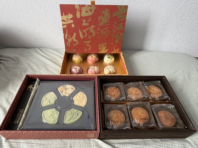 A variety box of gourmet mooncakes is displayed on a bed, featuring an open red and gold ornamental box with floral-decorated pieces on top, alongside two trays containing intricately molded traditional mooncakes. The packaging's golden Chinese calligraphy and elegant presentation suggest a premium gift, fitting the user's caption about receiving a single box as a KOL gift.
