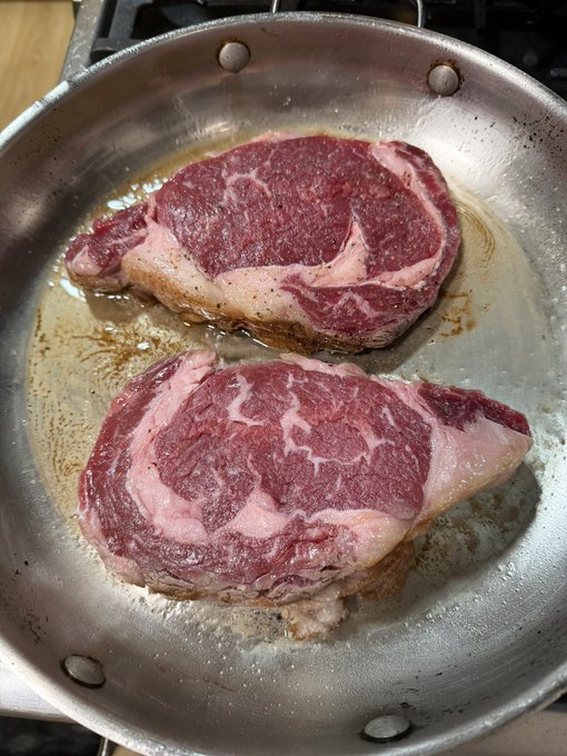 Two thick, raw steaks are being seared in a stainless steel pan, showing nice marbling and browned edges.