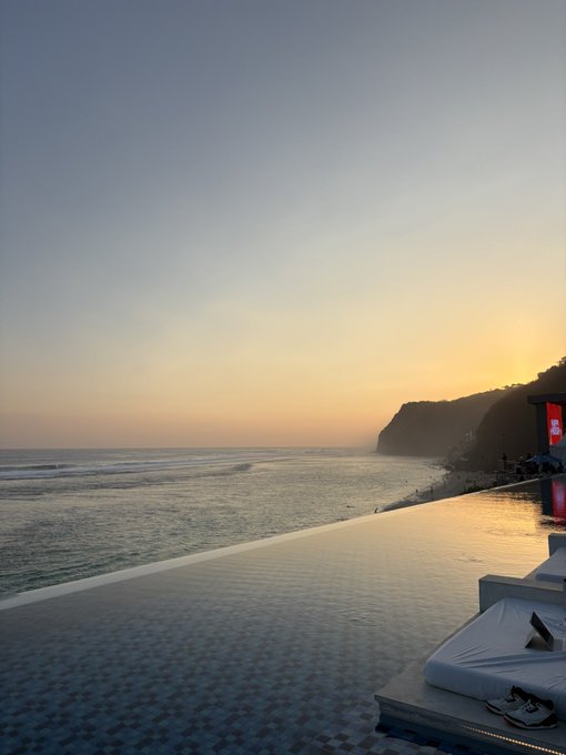A tranquil beach scene at sunset with calm waves rolling onto a tiled infinity pool deck, complete with white loungers and a pair of sneakers, set against a warm coastal horizon. The serene "vibes" of the scene serve as a visual metaphor for the chill, lifestyle-oriented ethos of trading Base ecosystem assets.