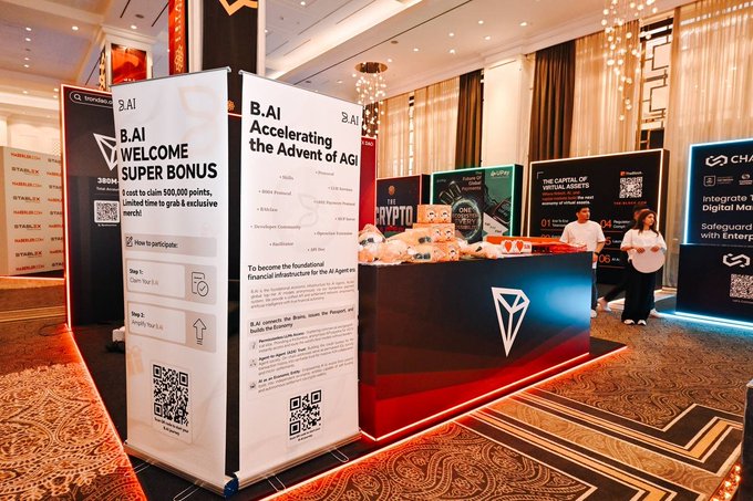 The image shows a booth with a red background, white text, and various information displayed for an event, likely a conference or exhibition. A man and a woman are standing near the booth, possibly attending to visitors or discussing the exhibit. Other people can also be seen around the area, some closer to the booth and others further away. 

In addition to the booth-goers, there is a laptop placed on one of the stands, which might be used for displaying information or controlling event aspects. The setting suggests that this gathering may be related to B.AI, super bonus, or cryptocurrency topics, given the presence of informational displays and people discussing such matters.